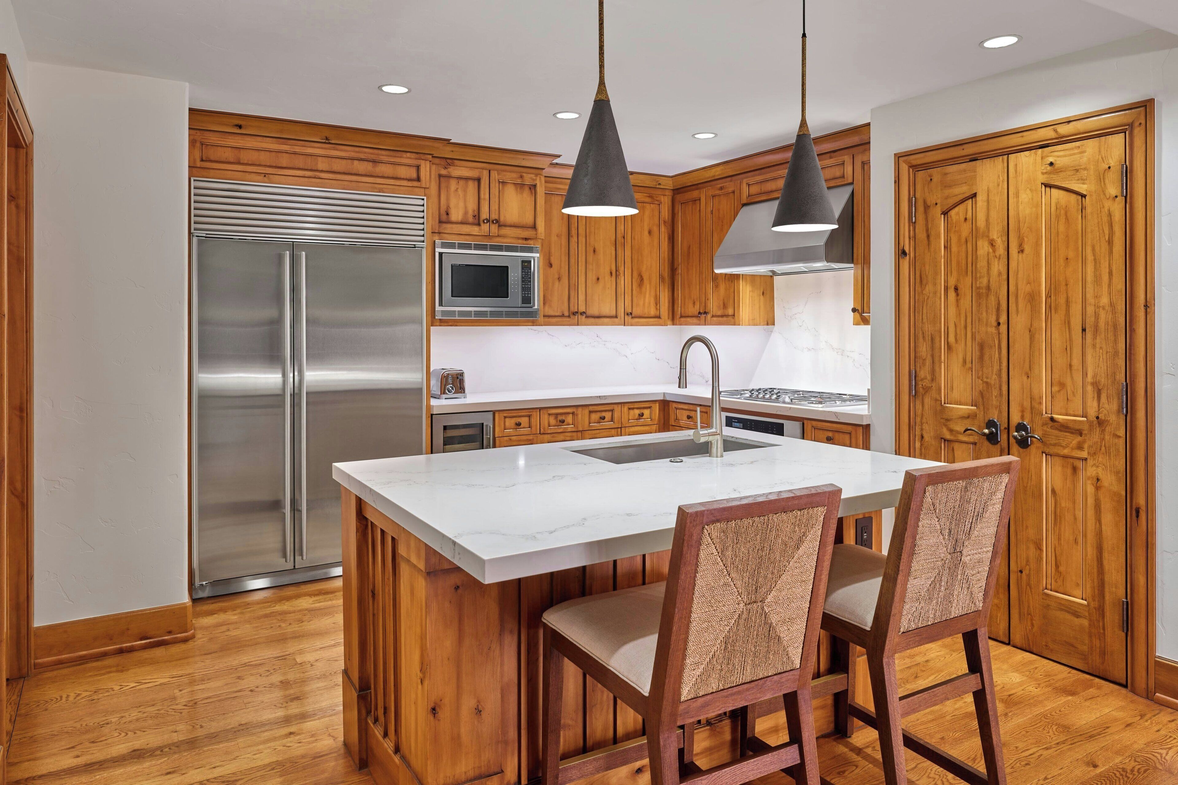 A kitchen with wooden cabinets, a stainless steel refrigerator, a microwave, and a sink. There are two bar stools with woven seats.