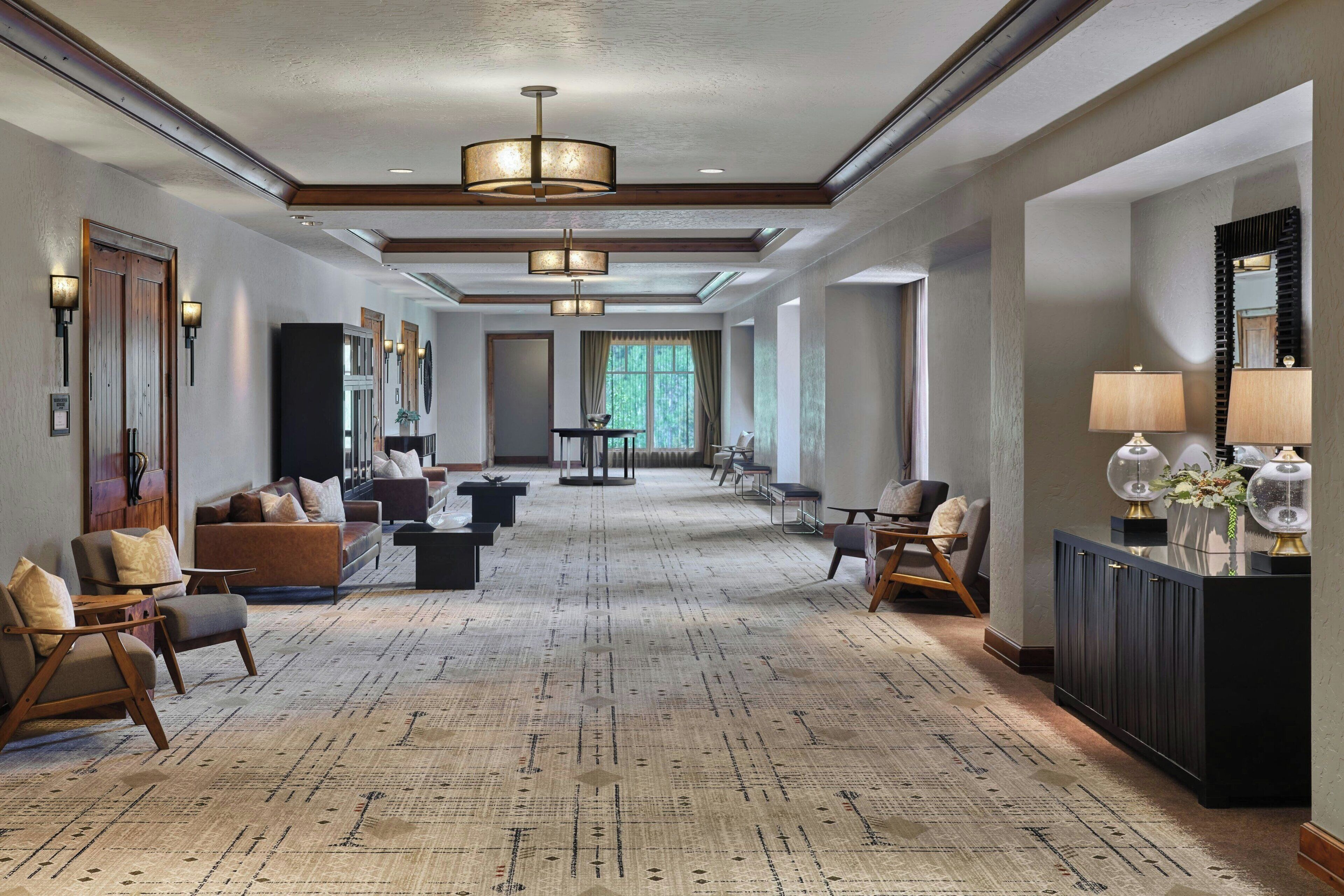 A spacious hotel lobby with a patterned carpet, multiple seating areas, and a large window providing natural light.