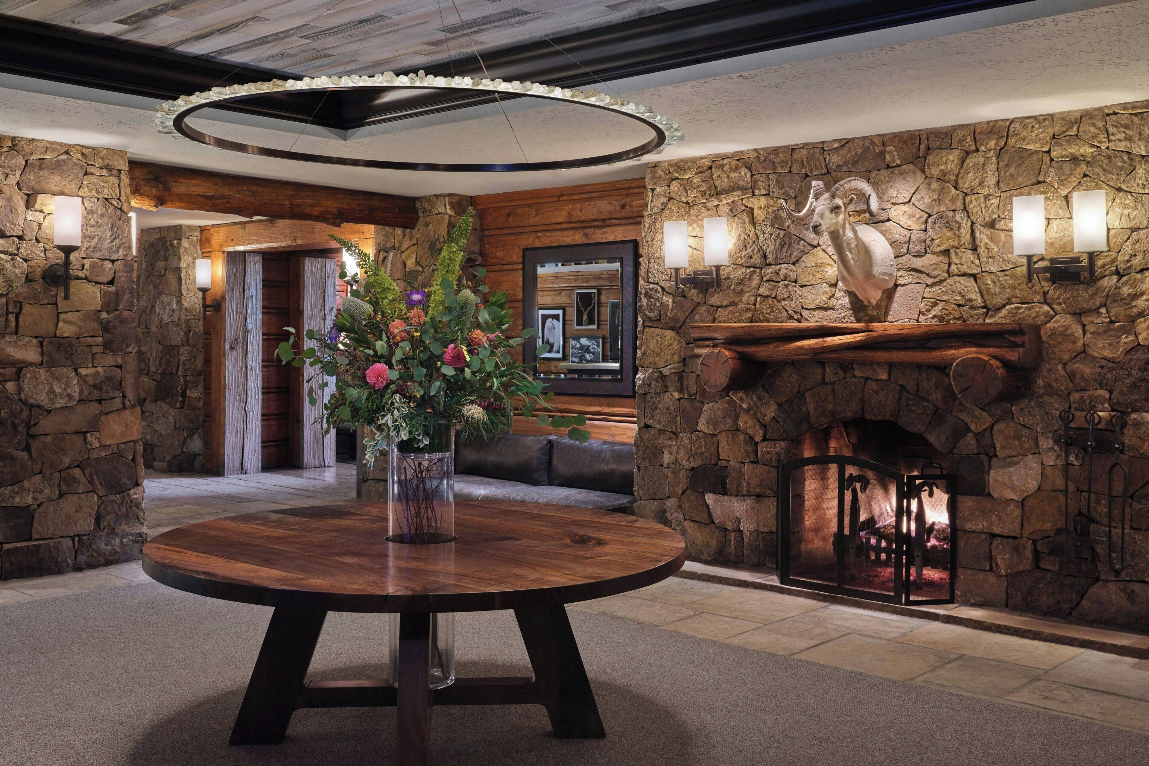 A lobby with a stone fireplace, a round wooden table with a vase of flowers, and a stone wall with a ram sculpture.