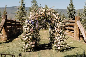 A rustic wooden archway adorned with white and purple flowers, set against a backdrop of mountains and trees.