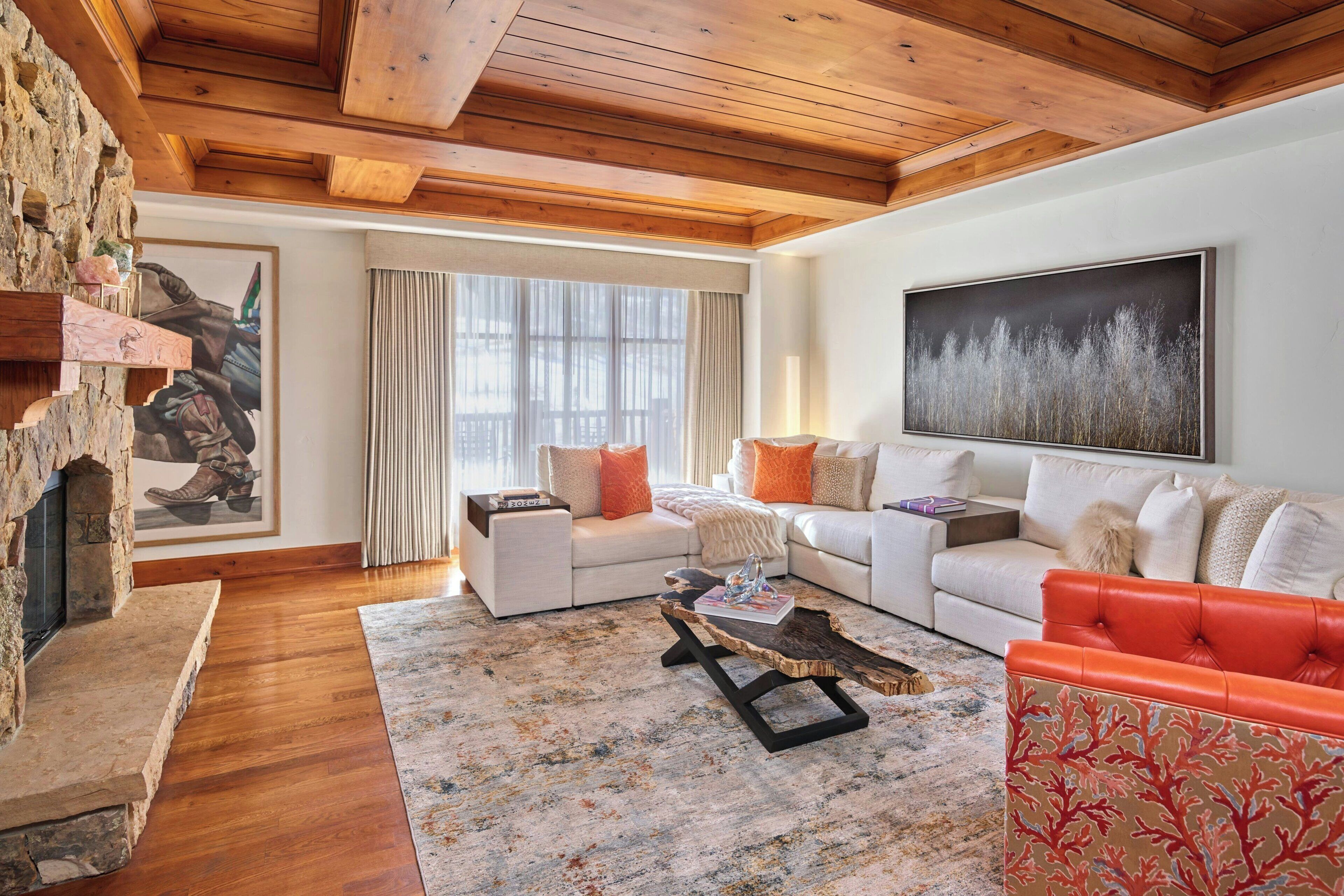 A living room with a stone fireplace, two white sofas with orange cushions, a red sofa, a wooden coffee table, and a large painting on the wall.