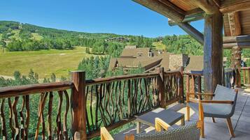 A wooden balcony with wicker furniture overlooking a green landscape and buildings.