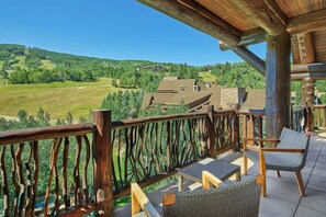 A wooden balcony with wicker furniture overlooking a green landscape and buildings.