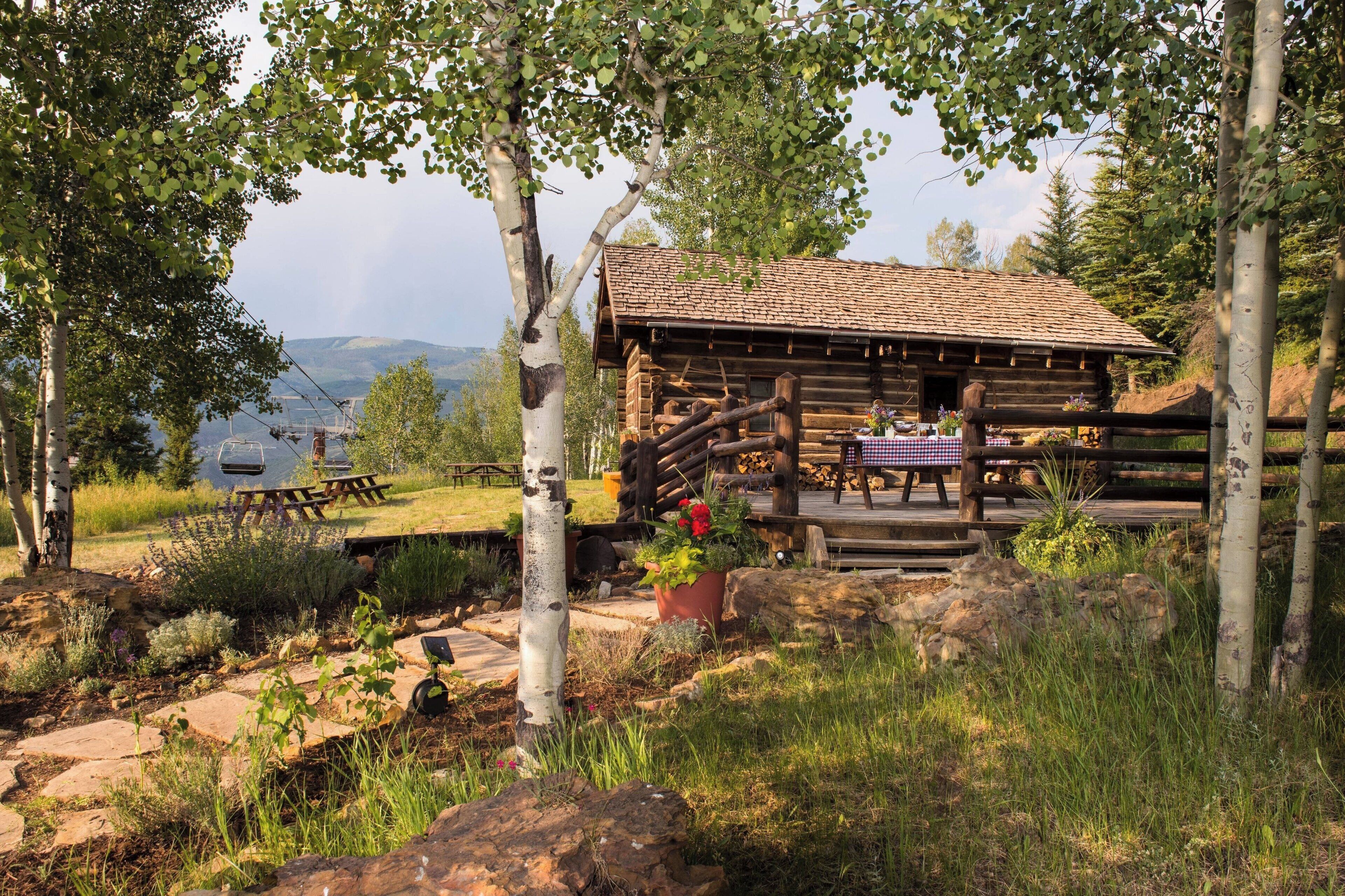 En rustik stuga med en träveranda, omgiven av träd och ett picknickbord.