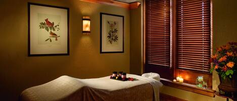 A room with a massage table, framed botanical prints on the wall, a window with blinds, and a vase with flowers on a small table.