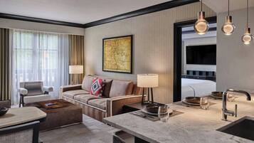 Een moderne hotelkamer met een bank, fauteuil, eettafel en een groot raam met gordijnen.