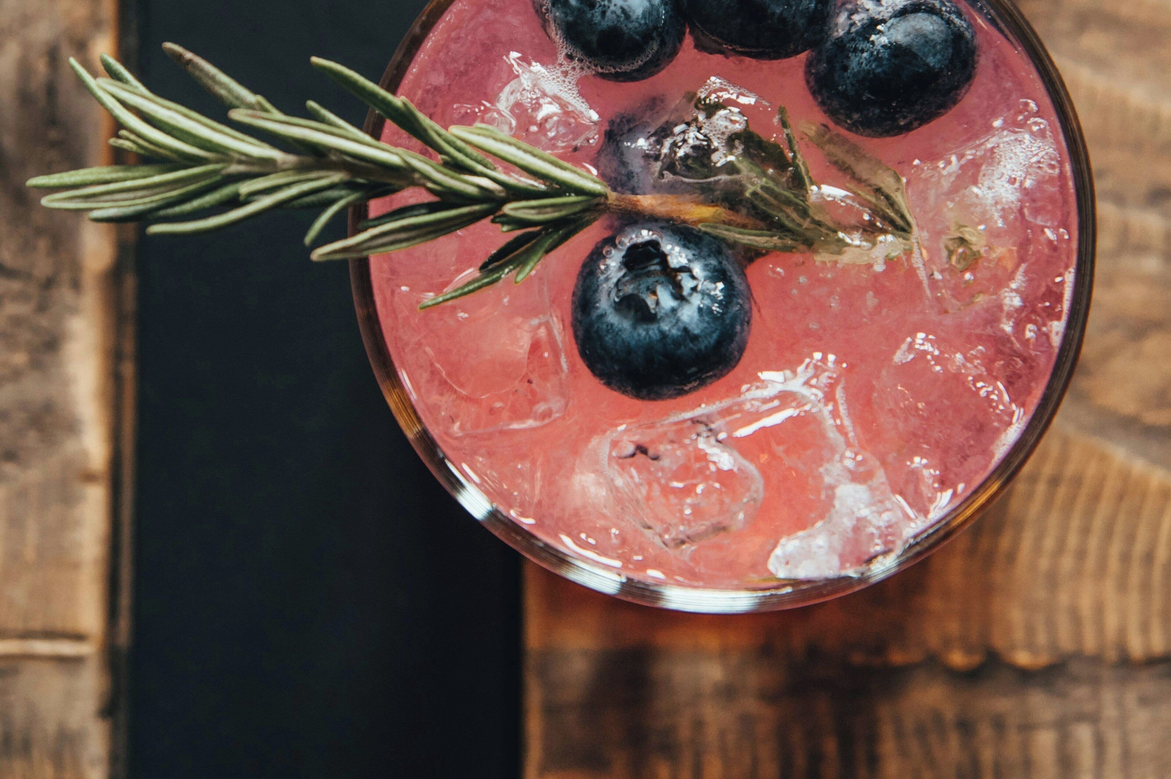 A glass of red beverage with ice, blueberries, and a sprig of rosemary.