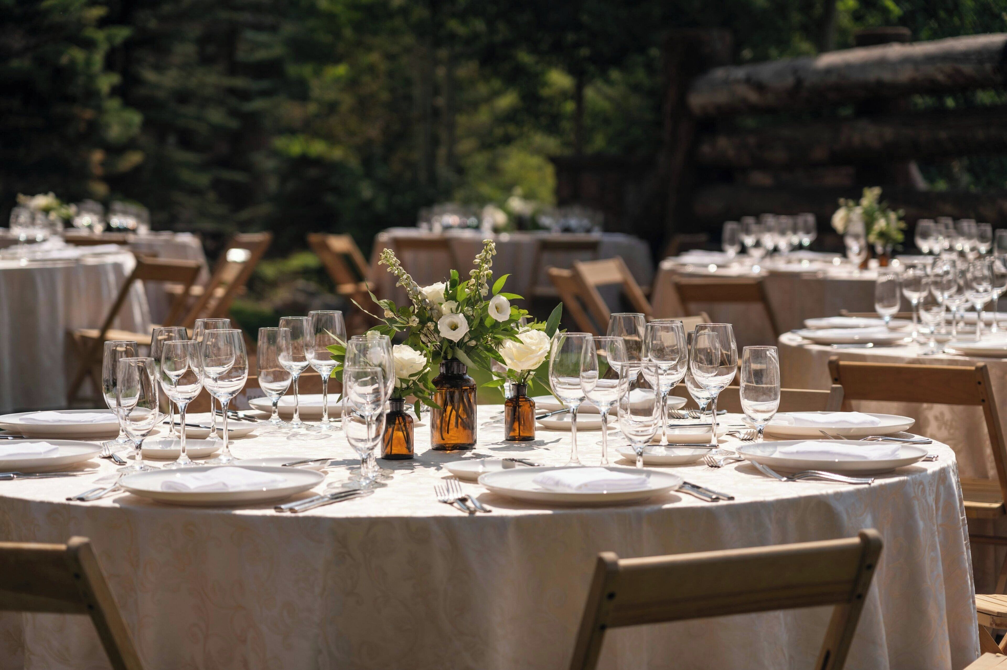 A series of round tables set outdoors with white tablecloths, each with a centerpiece of white flowers and a small bottle.