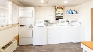 A laundry room with four washing machines, a dryer, a wall-mounted laundry bar, and a shelf with detergent bottles. - Circle S Motel Cozad I-80 (Cozad)