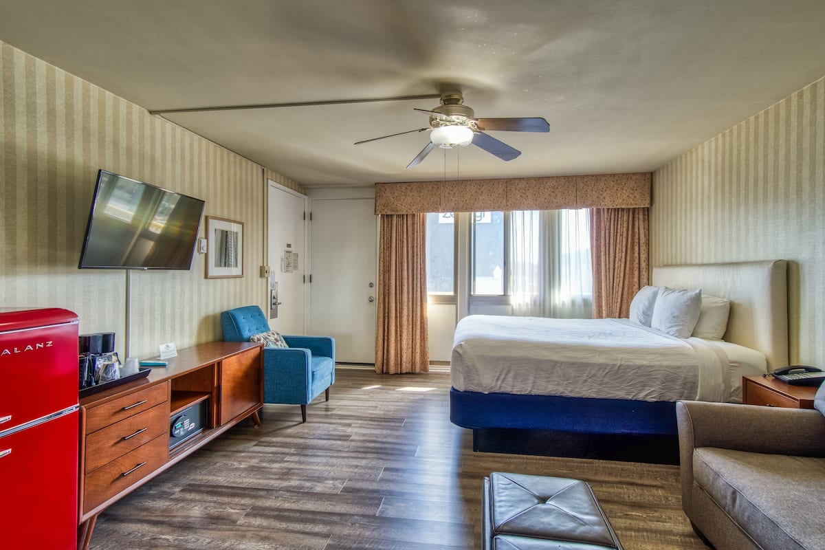 A hotel room with a bed, a TV, a red minibar, a blue armchair, and a ceiling fan.