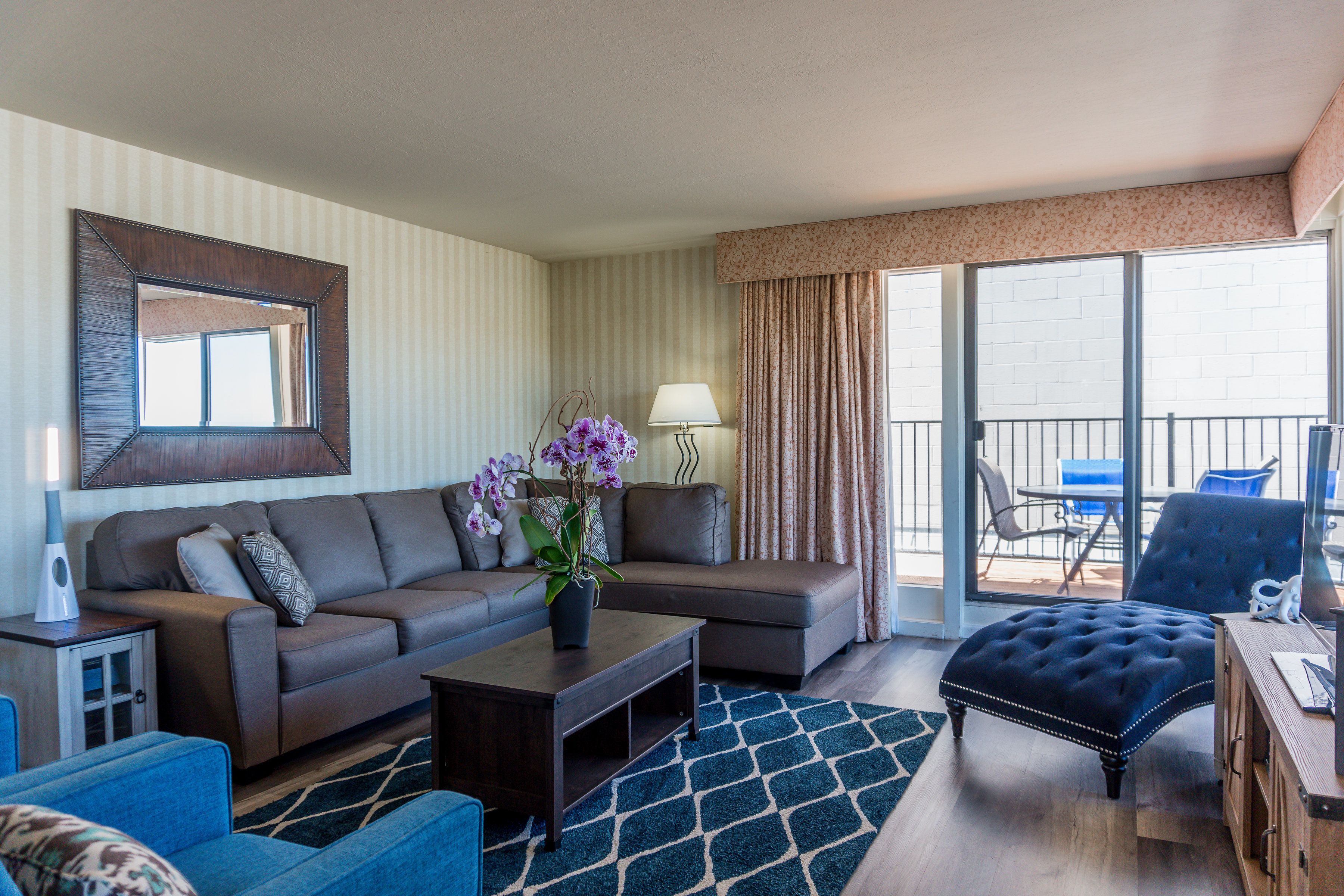 A living room with a sofa, ottoman, coffee table, and a lamp. There is a balcony with chairs and a table.
