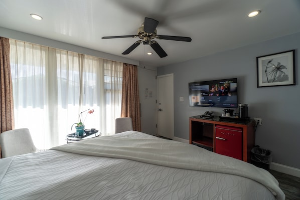 A hotel room with a bed, a ceiling fan, a TV, a desk with a coffee maker, and a red cabinet.