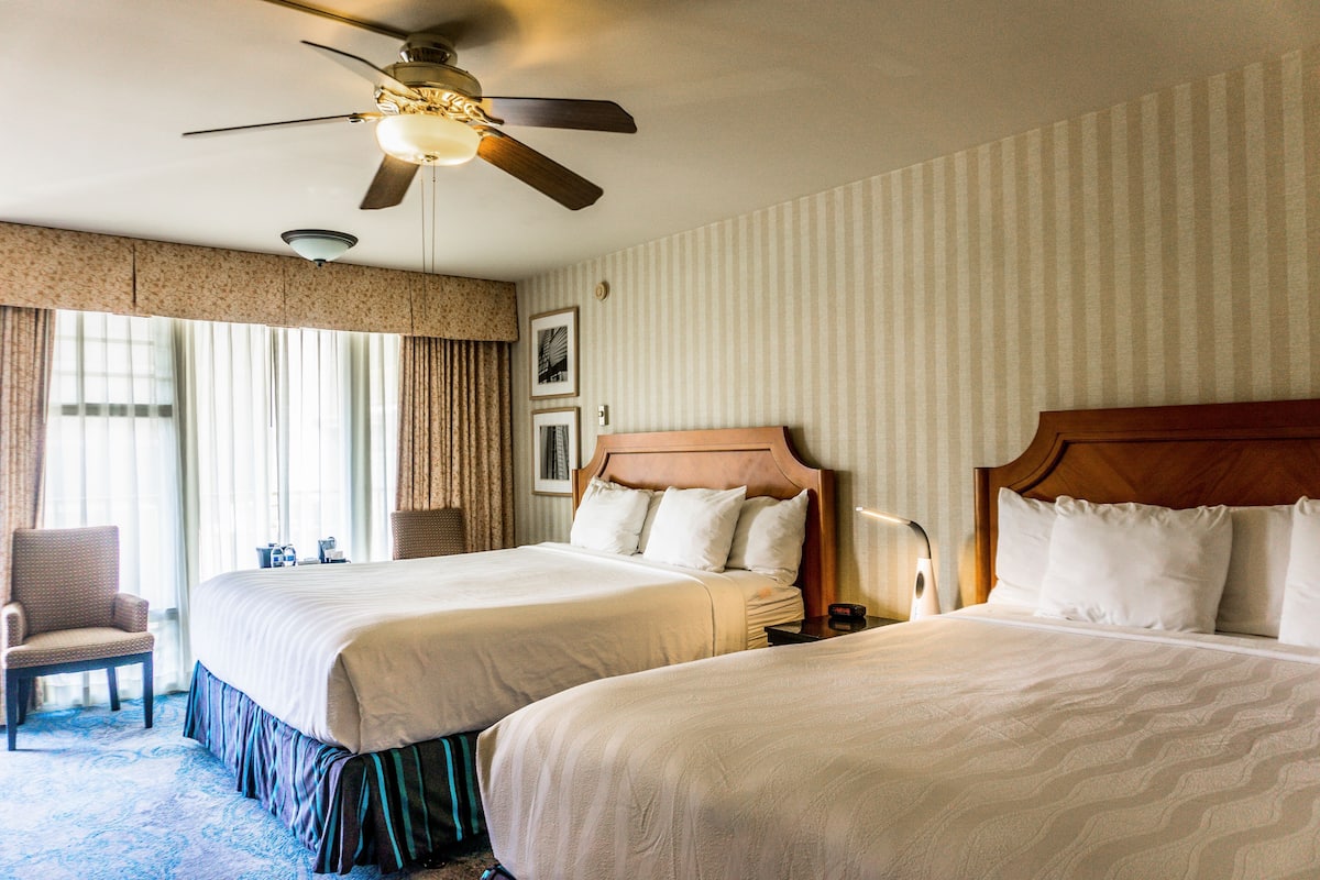 A hotel room with two beds, a ceiling fan, a chair, and a large window with curtains.