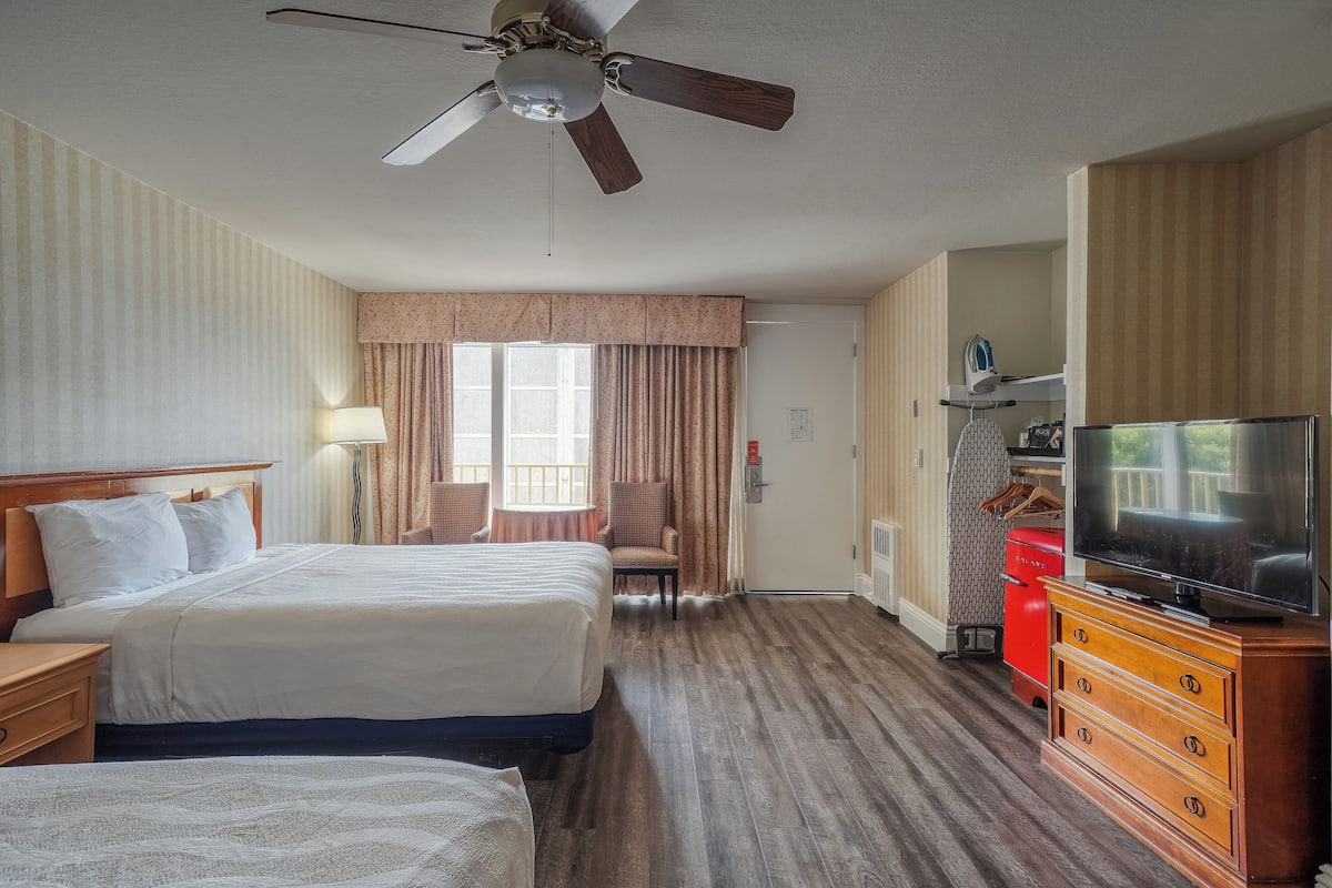 A hotel room with two beds, a ceiling fan, a television, a dresser, and a window with curtains.