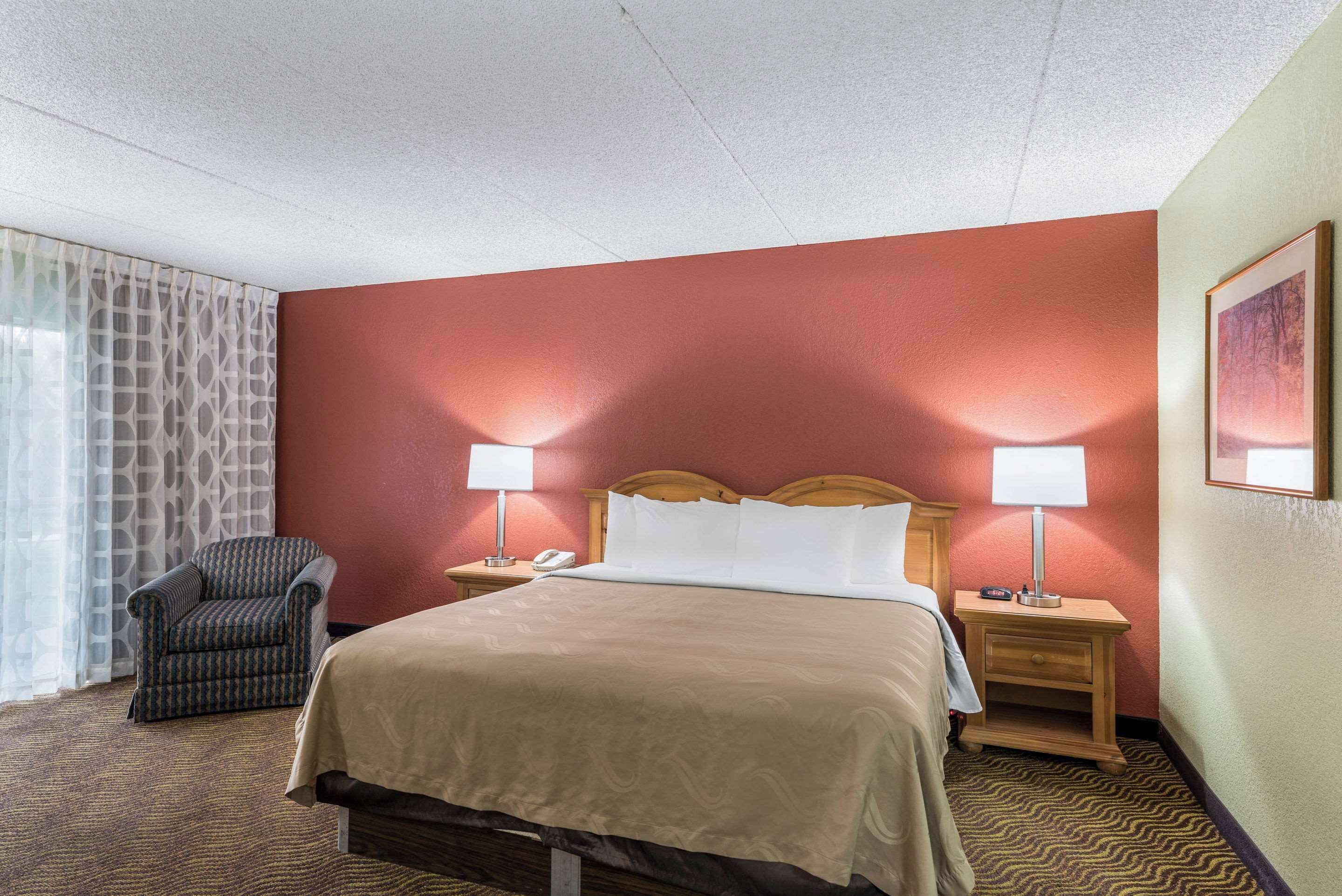 A hotel room with a bed, two bedside tables with lamps, a chair, and a window with patterned curtains.