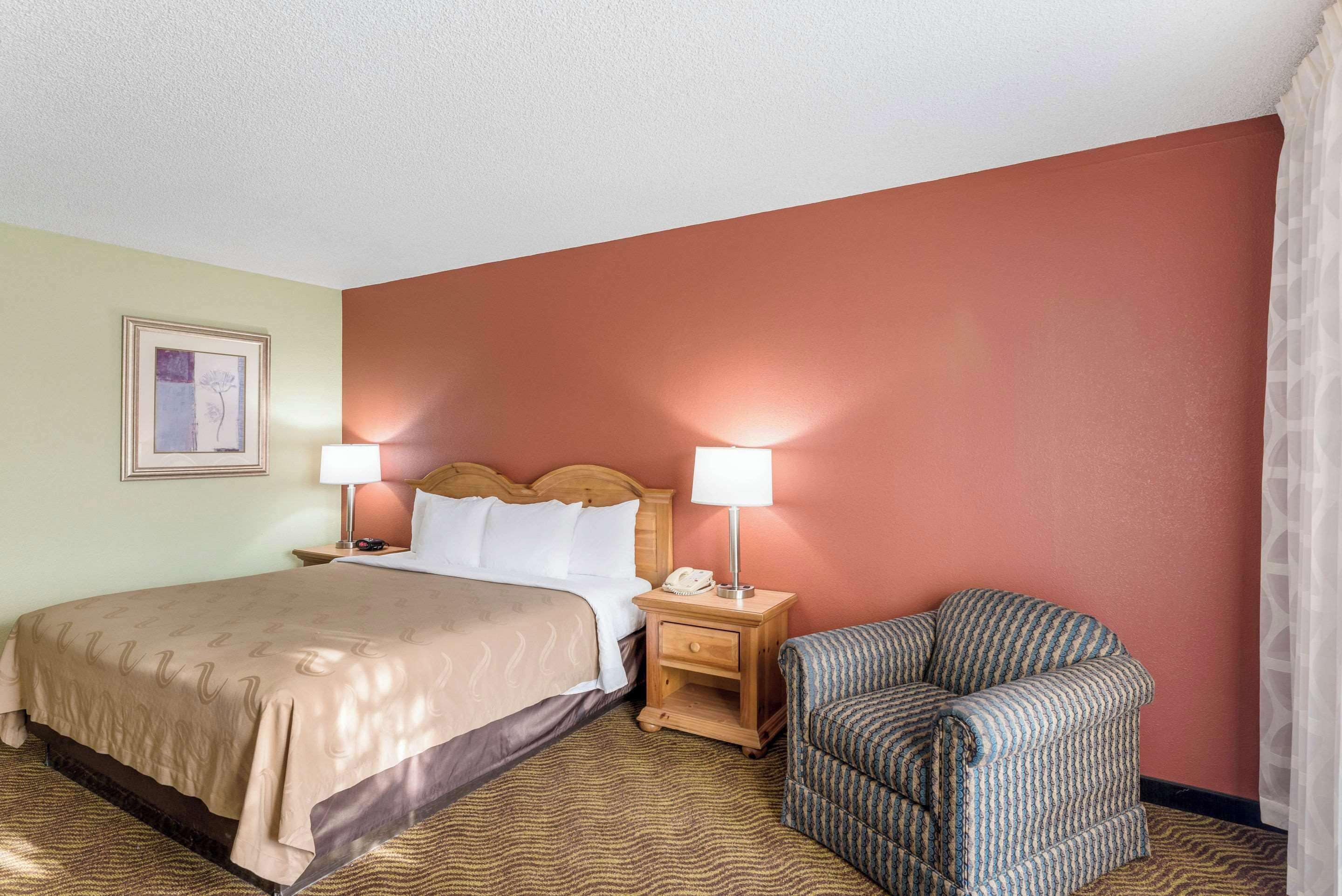 Una habitación de hotel con una cama, dos mesitas de noche con lámparas, una silla y un cuadro en la pared.