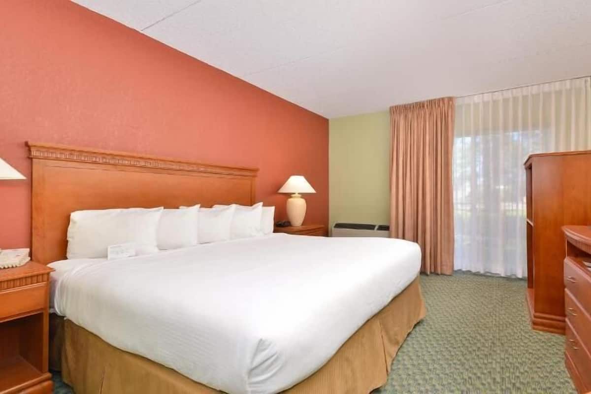 A hotel room with a large bed, a television on a wooden stand, a nightstand with a lamp, and a window with curtains.