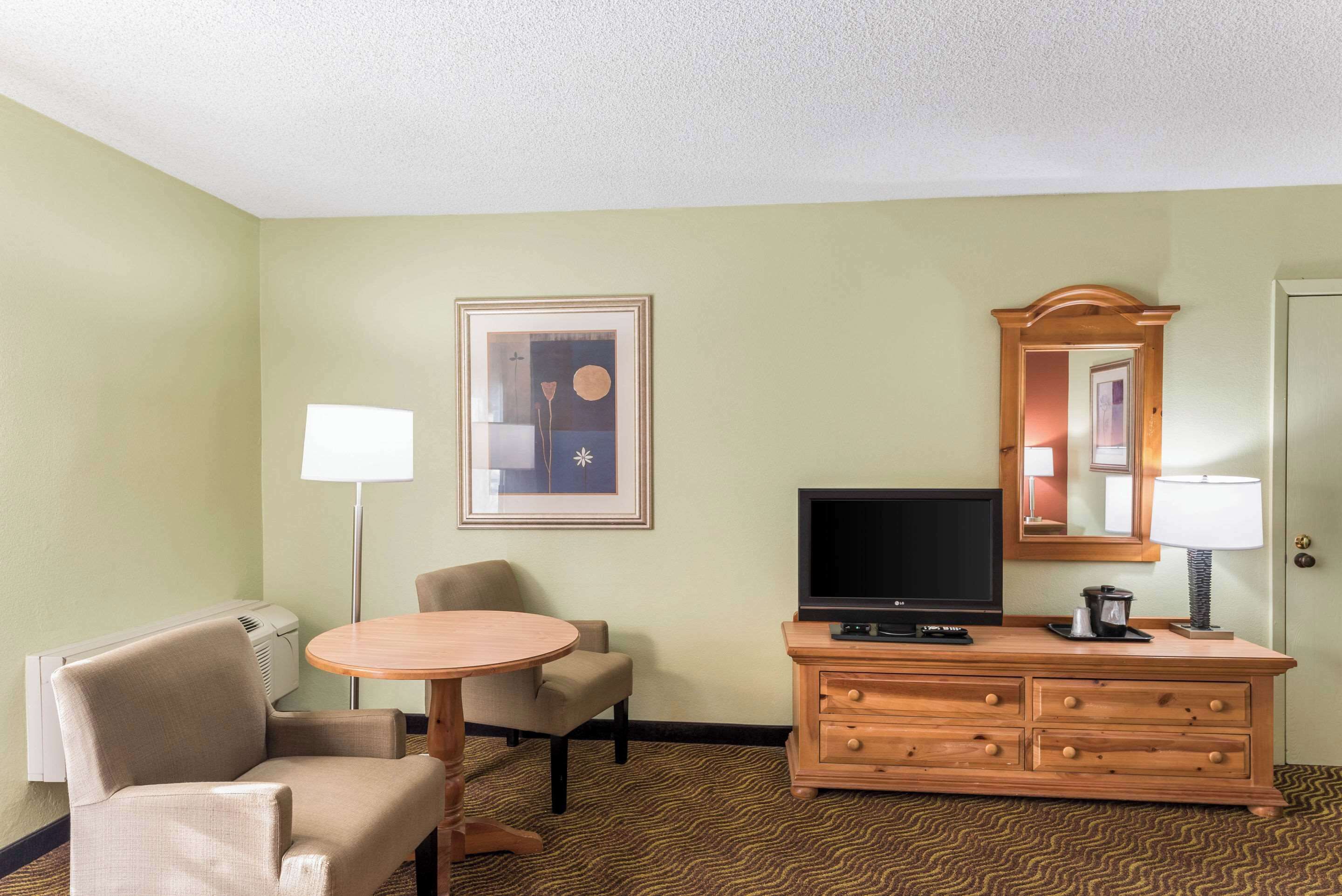 A hotel room with a round table, two chairs, a television, a dresser, a mirror, and a framed picture on the wall.