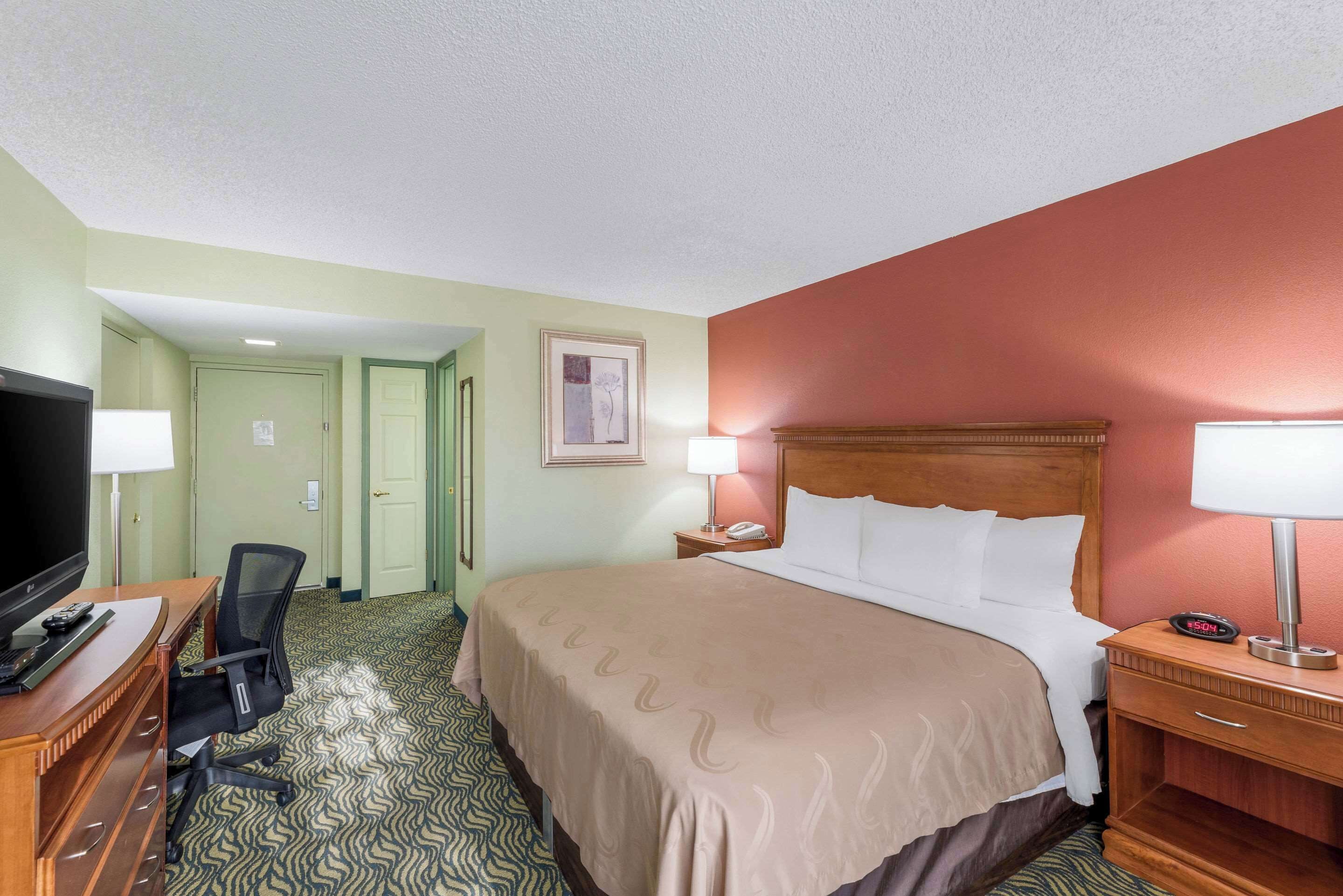 A hotel room with a bed, desk, chair, television, and a nightstand with a lamp.