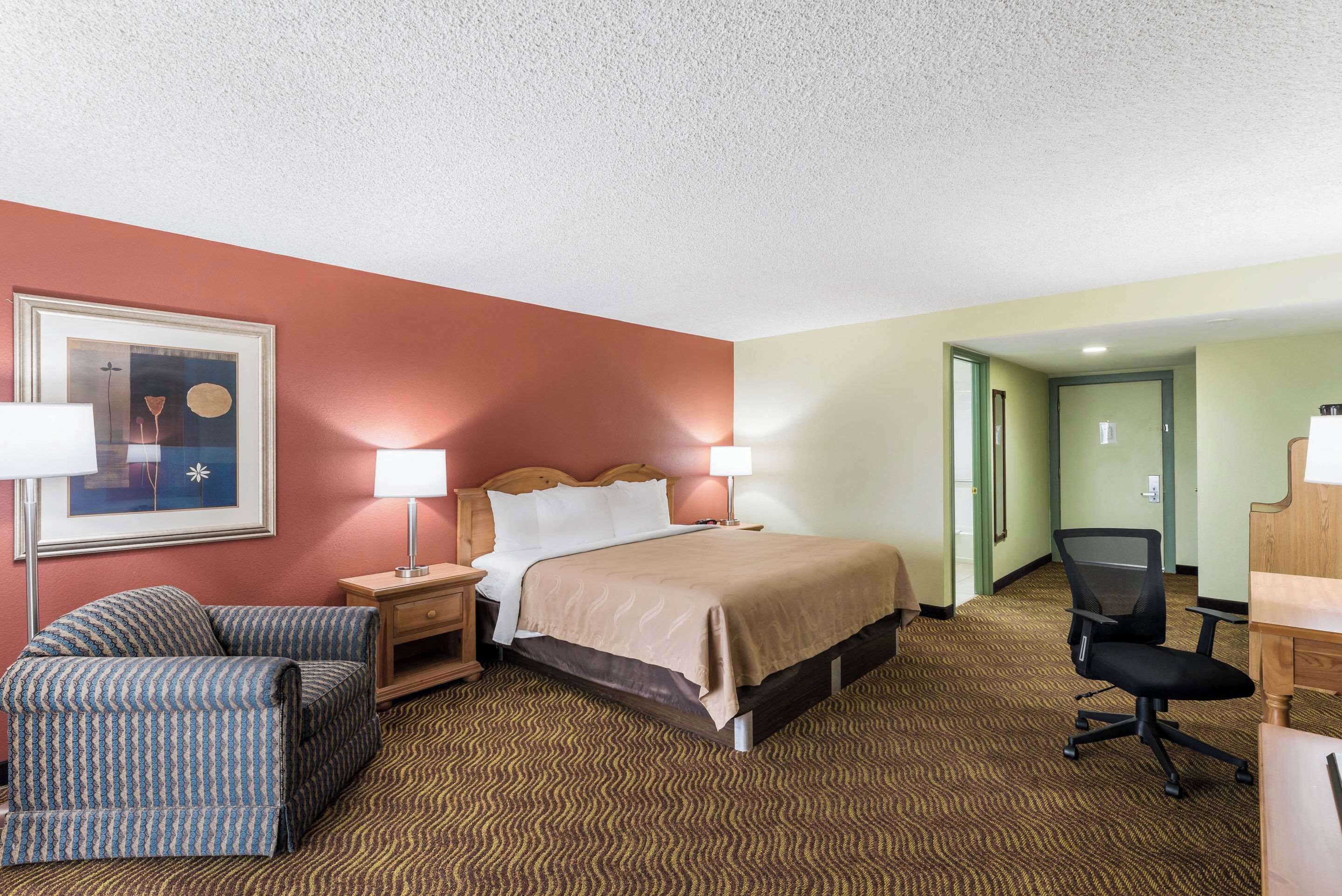 A hotel room with a bed, a chair, a desk, and a framed picture on the wall.