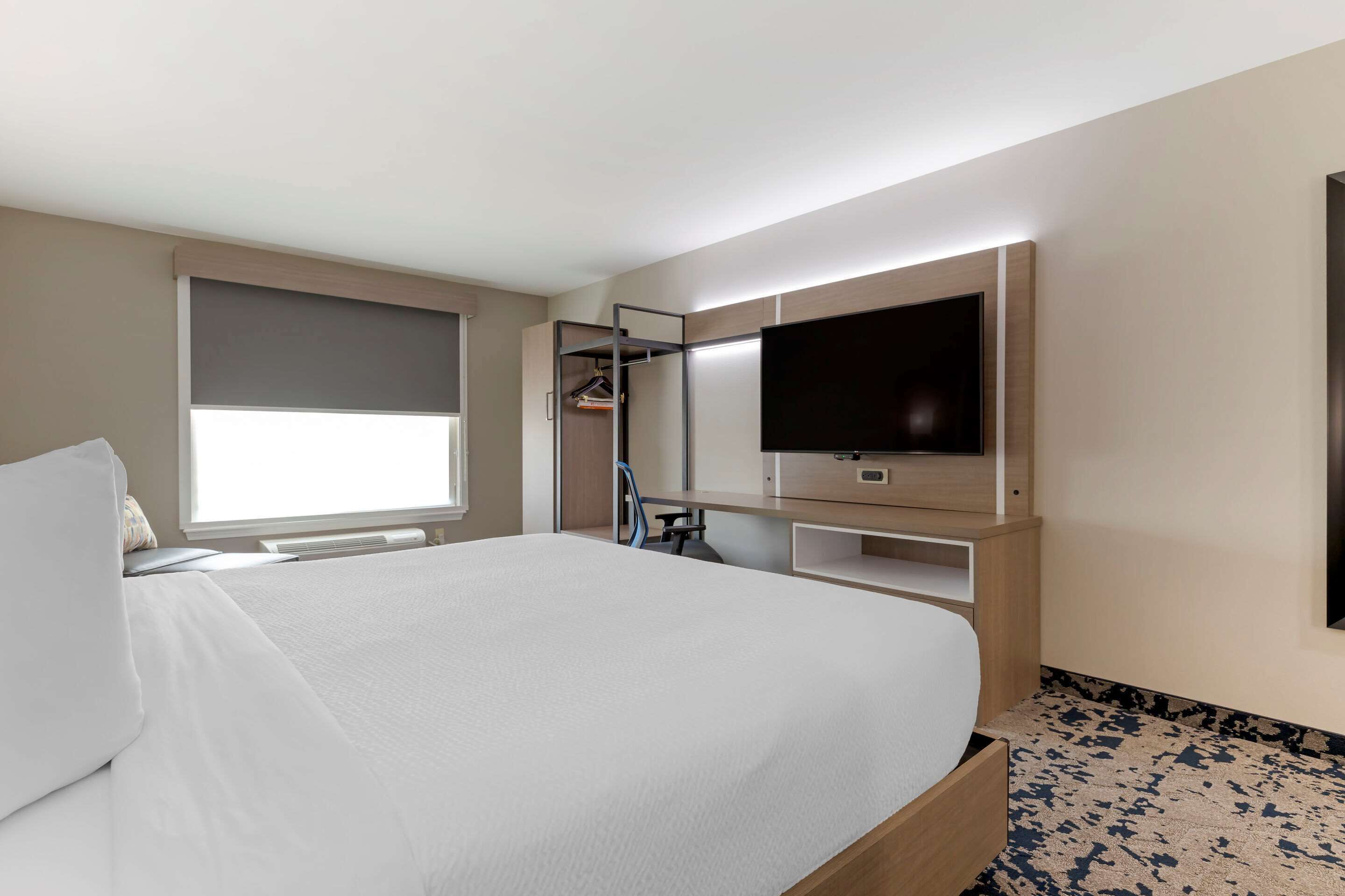 A modern hotel room with a large bed, a TV mounted on the wall, a wardrobe, and a window with blinds.