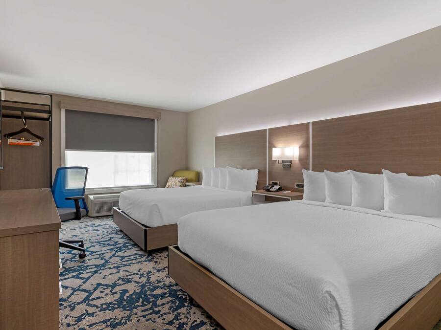 A hotel room with two beds, a desk, a chair, a window with curtains, and a wall-mounted lamp.