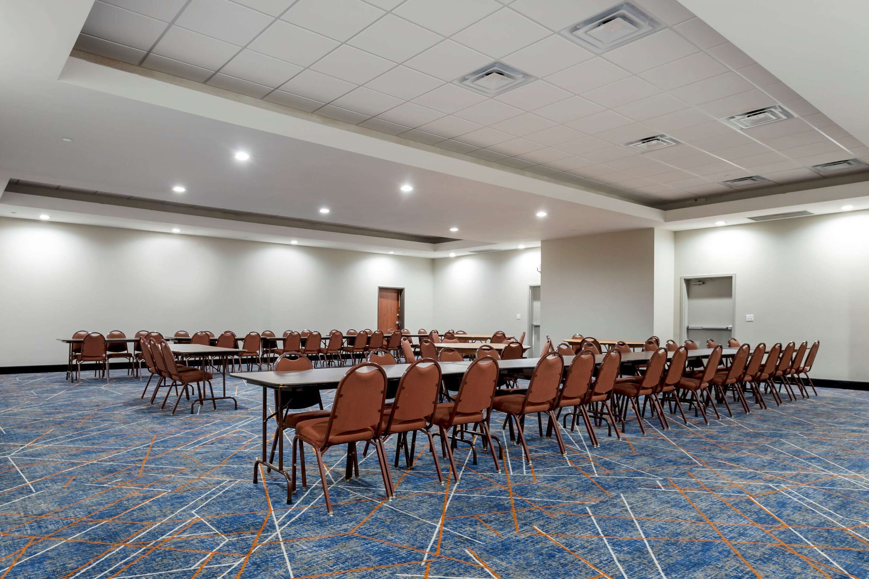 A conference room with rows of tables and chairs, a blue patterned carpet, and recessed lighting on the ceiling.