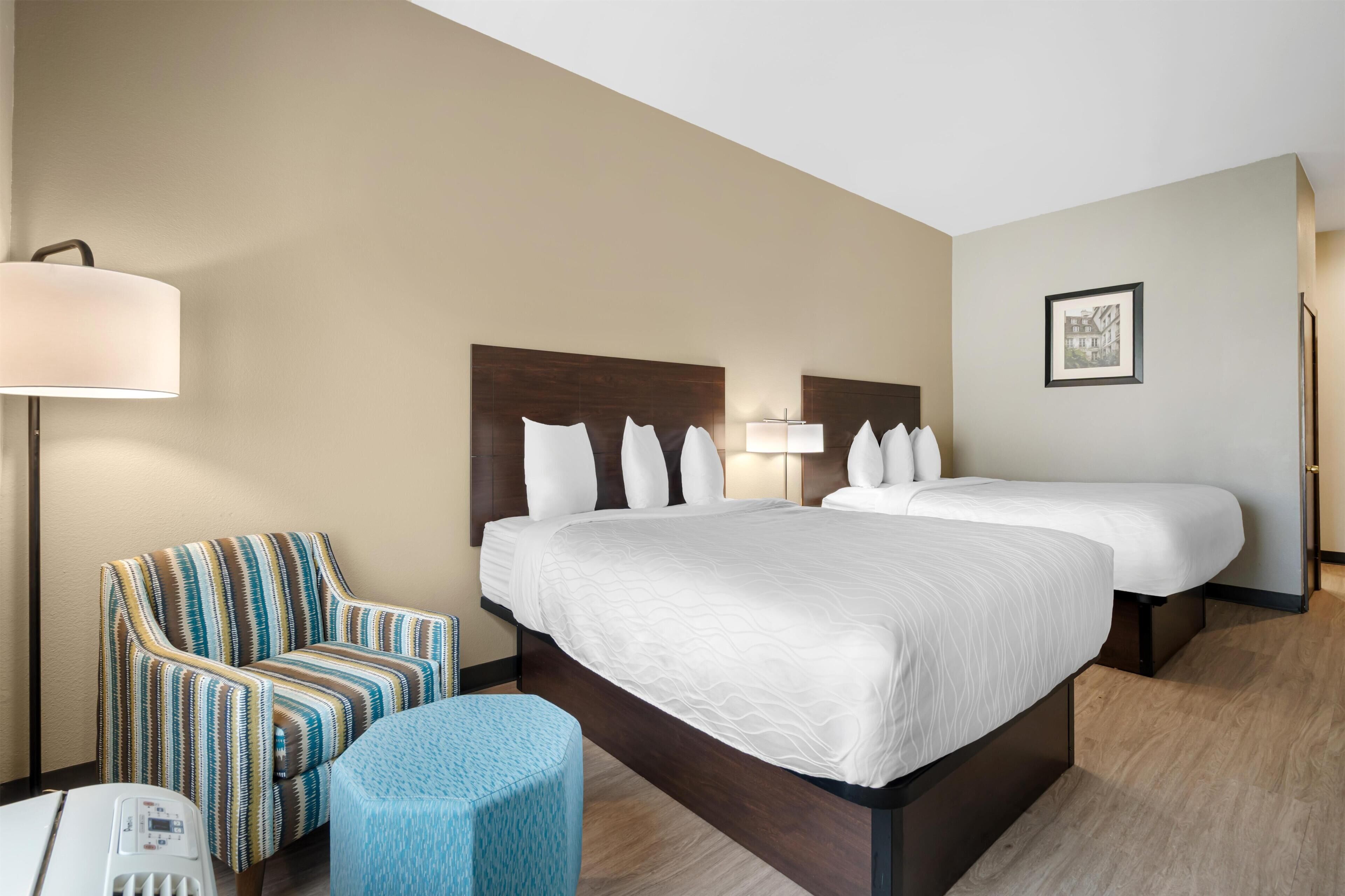 Una habitación de hotel con una cama grande, un sillón a rayas, un taburete redondo pequeño, una lámpara y un cuadro en la pared.