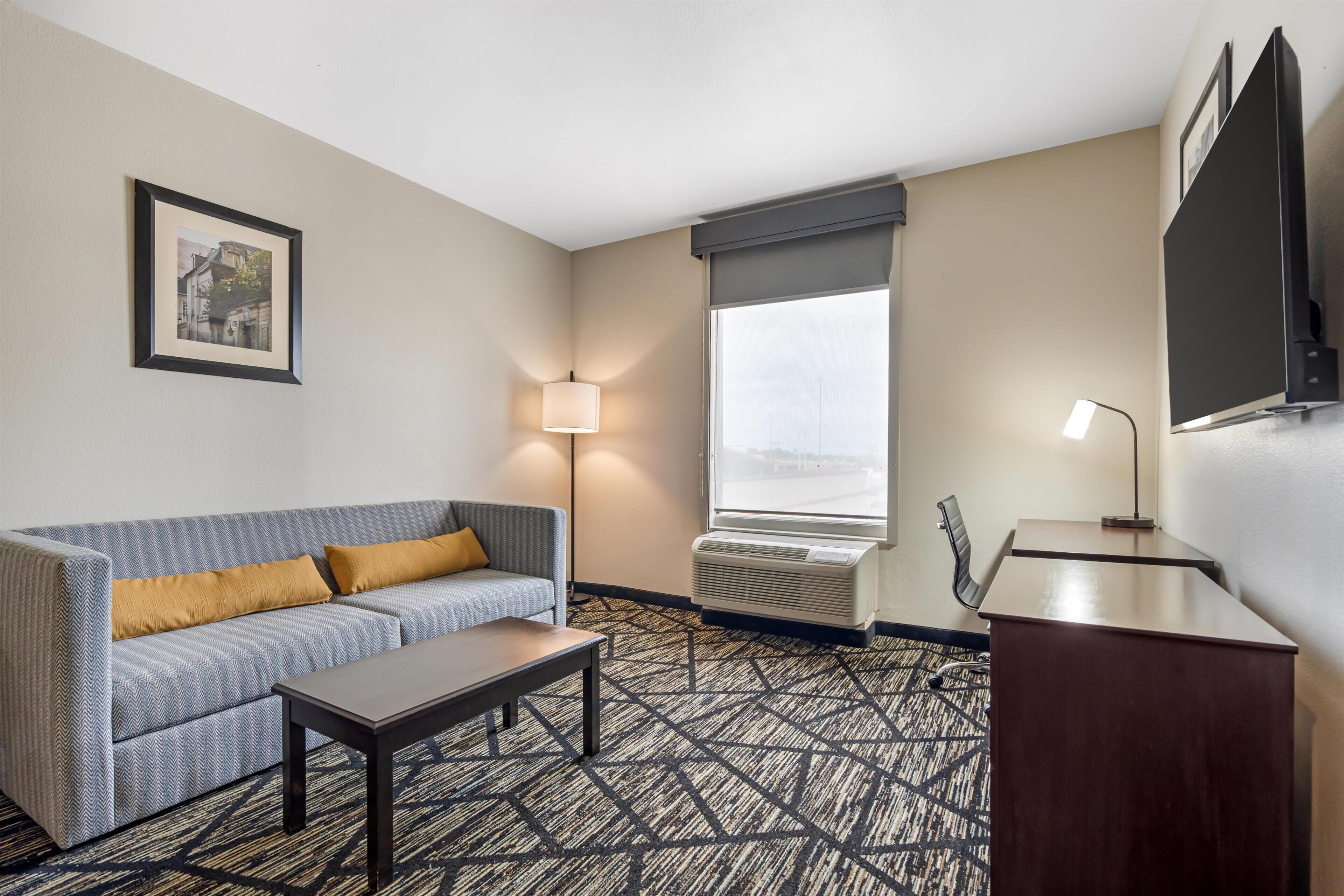 A hotel room with a sofa, a small table, a desk, a lamp, an air conditioning unit, a window with a view, and a framed picture on the wall.