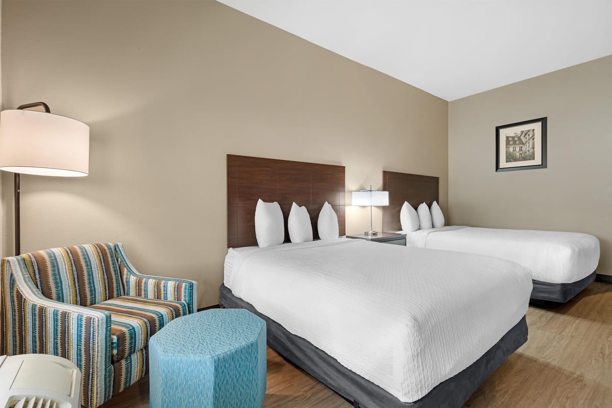 A hotel room with two beds, a striped armchair, a small round table, and a lamp.