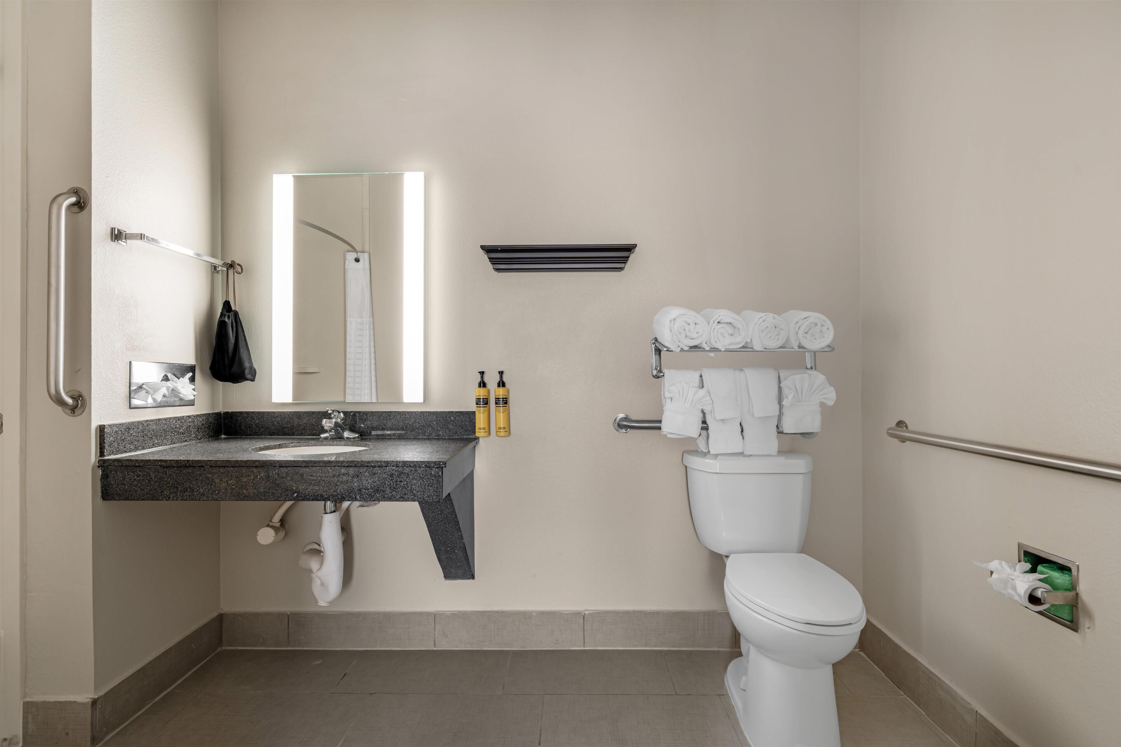 Un baño con lavamanos, espejo, inodoro, barras de apoyo y toallas.
