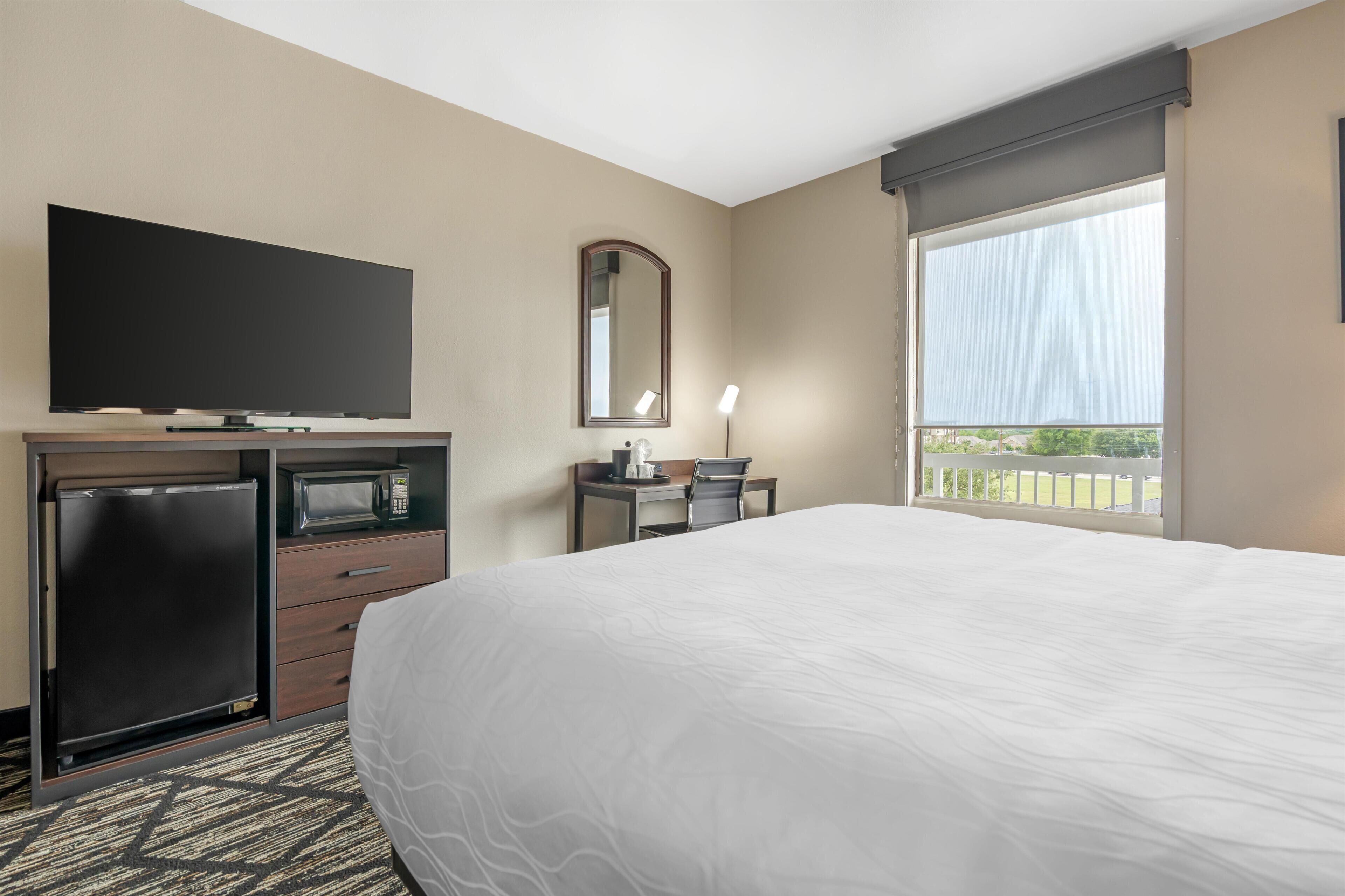 A hotel room with a bed, a flat-screen TV, a microwave, a refrigerator, a desk with a chair, a lamp, and a large window with a view.