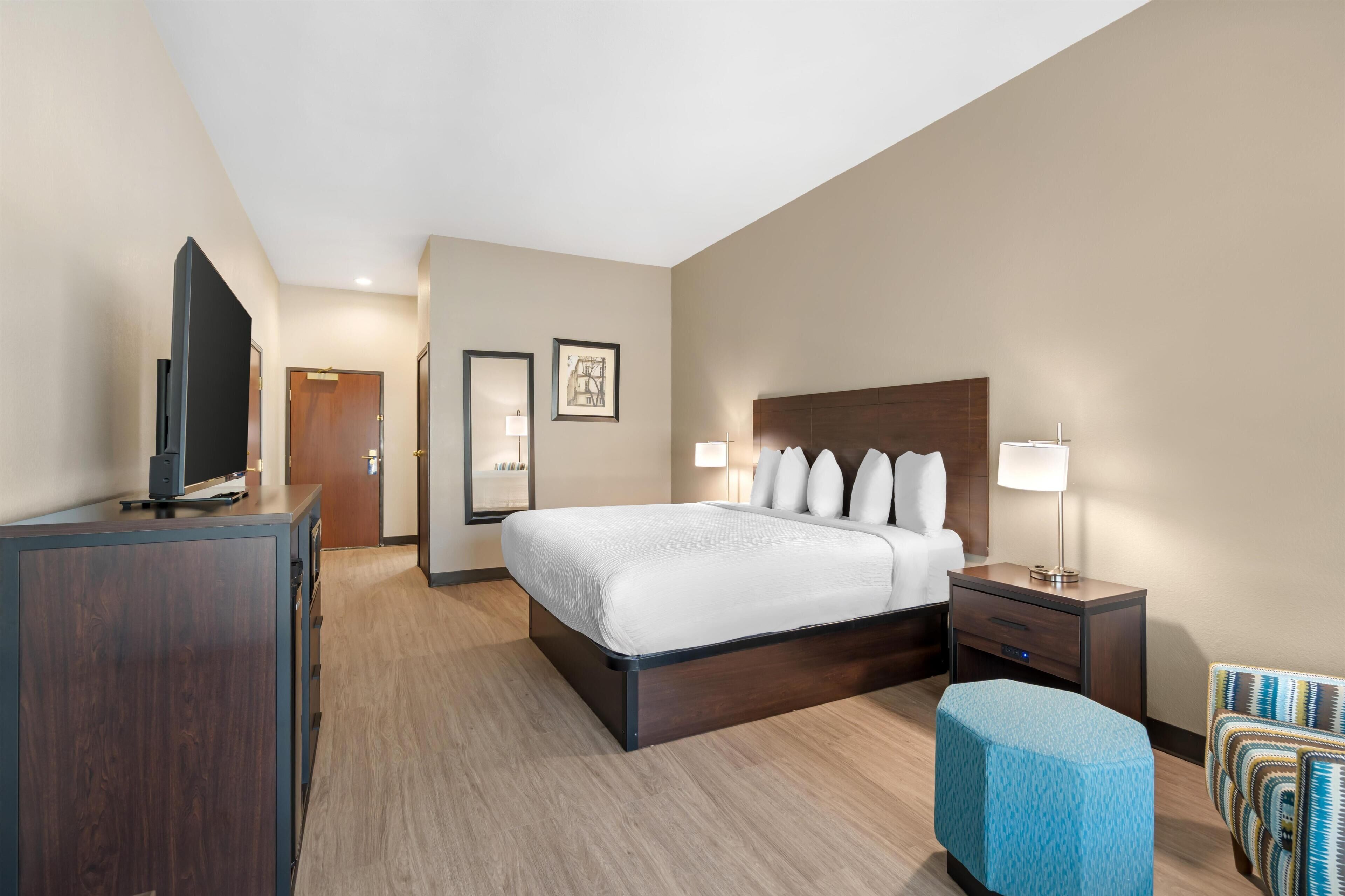 A hotel room with a bed, bedside table, lamp, TV, and a blue ottoman.