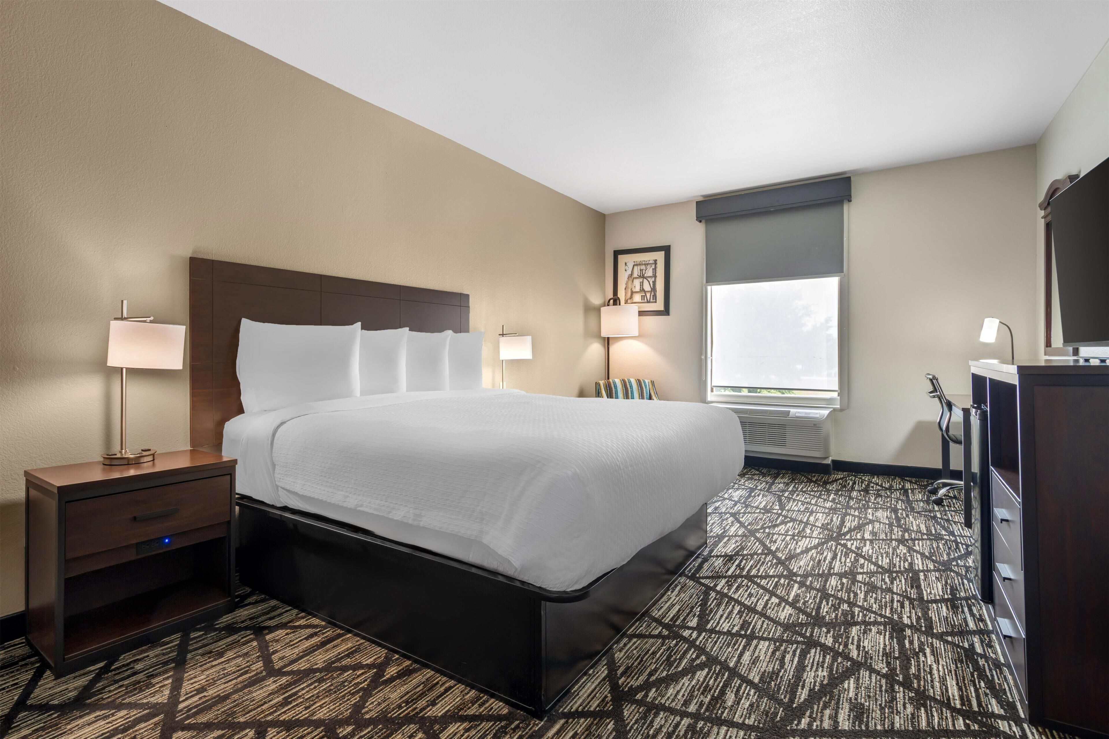 A hotel room with a bed, bedside tables, lamps, a desk, and a window.