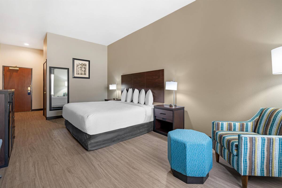 A hotel room with a bed, bedside tables, a chair, a small ottoman, and a mirror.