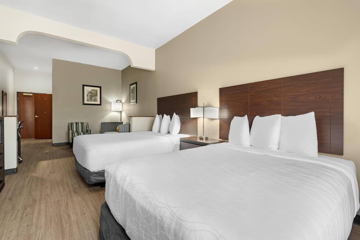 A hotel room with two beds, a wooden headboard, a desk, a chair, a lamp, and a door.