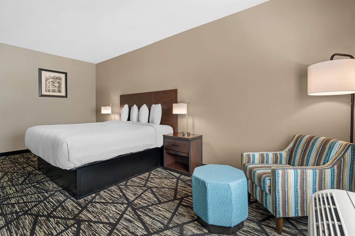 A hotel room with a bed, bedside table, lamp, chair, and ottoman.