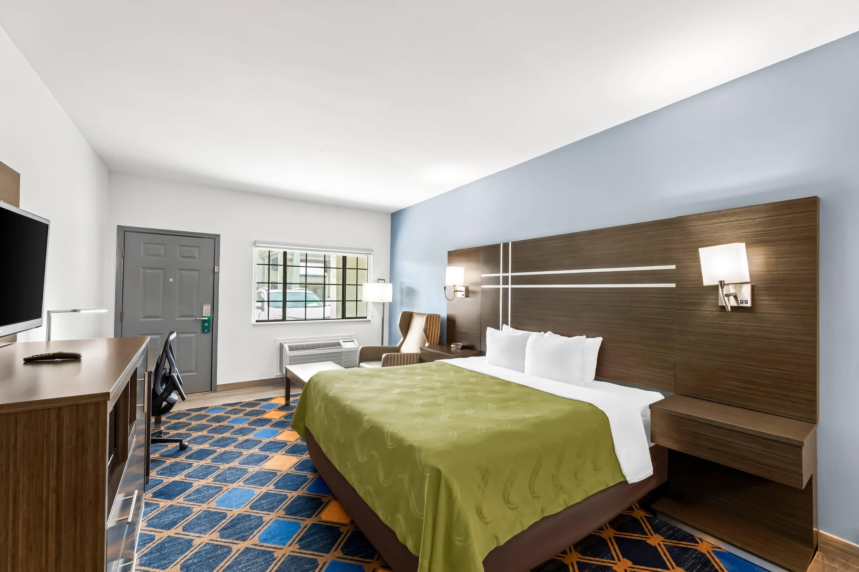 Quarto de hotel com cama, escrivaninha, cadeira, televisão e janela.