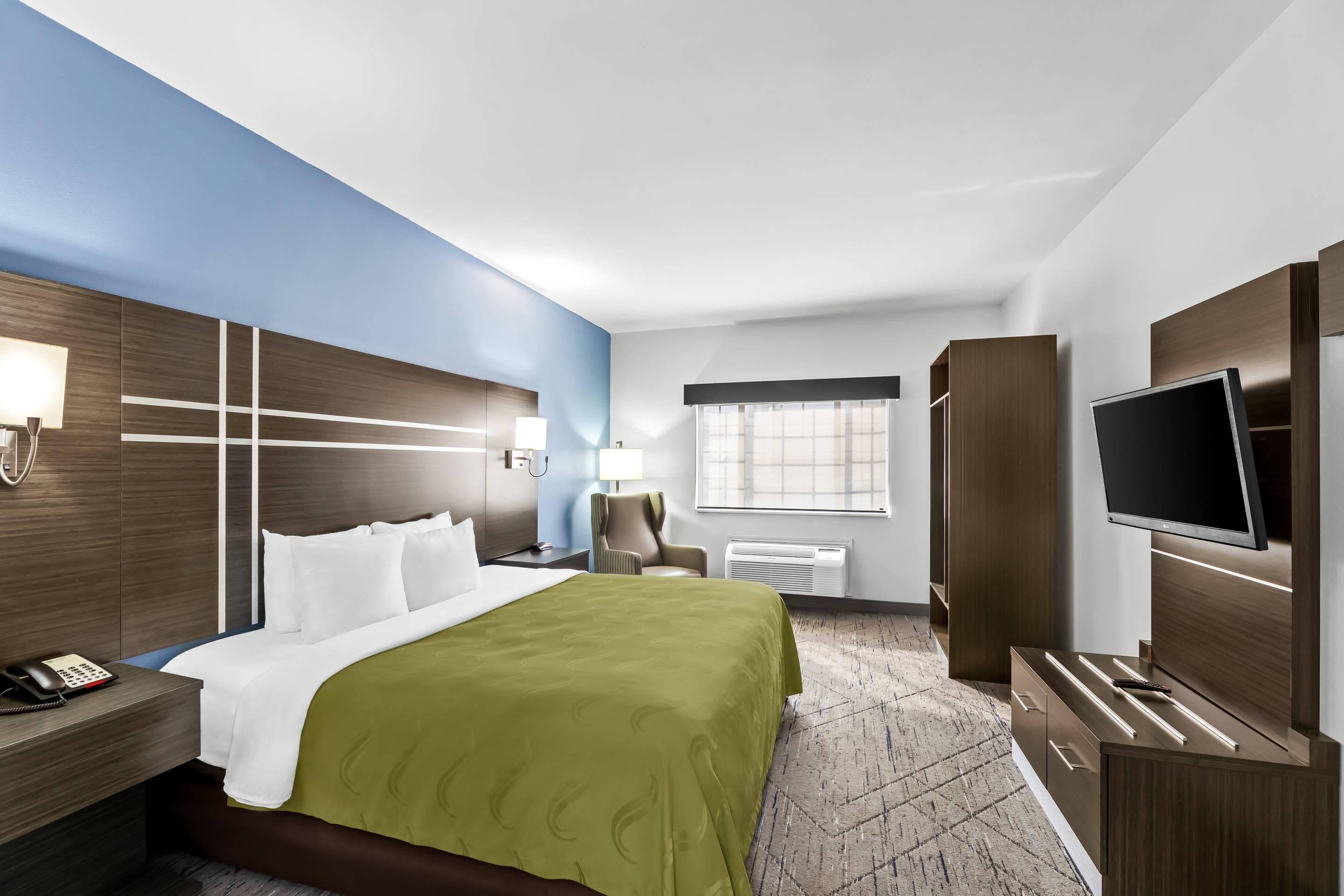 A hotel room with a bed, bedside table, lamp, chair, TV, wardrobe, and a window with blinds.