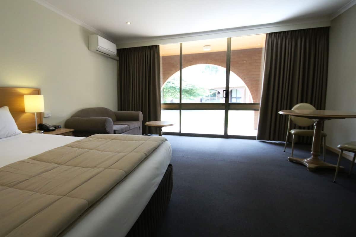 A hotel room with a large bed, a desk, a chair, and a small table. There is a window with curtains and a view of the outside.