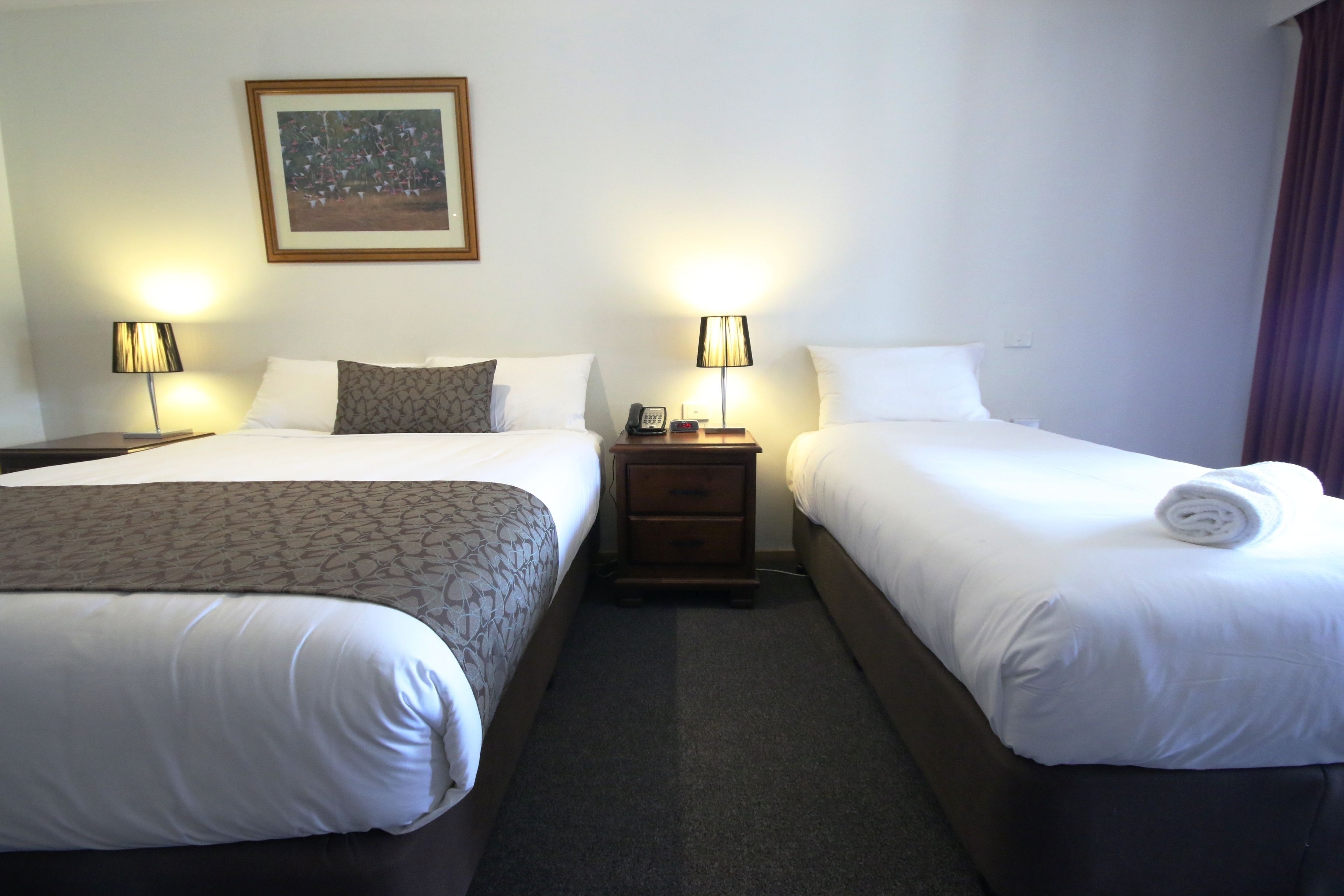 Une chambre d’hôtel avec deux lits, chacun agrémenté de draps blancs et d’une couvre-lit à motifs. On y trouve des tables de chevet avec des lampes, un tableau encadré au mur et une serviette roulée sur l’un des lits.
