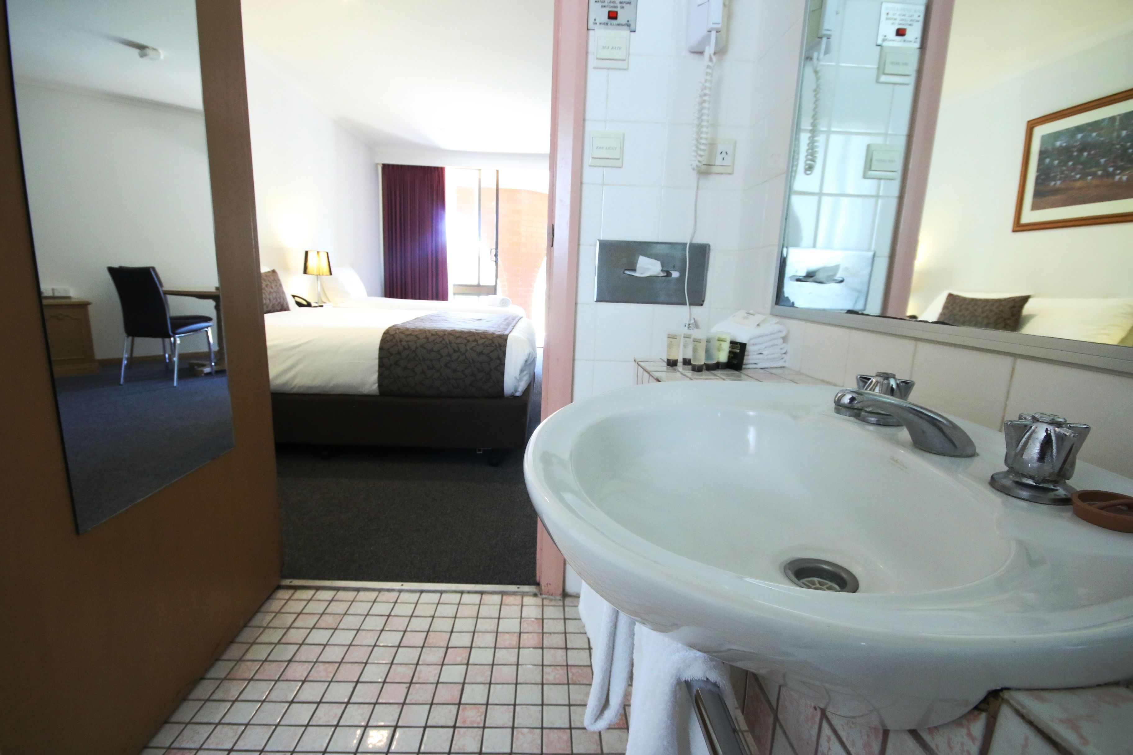 Habitación de hotel con una cama grande, un baño con lavamanos y espejo, y una silla.