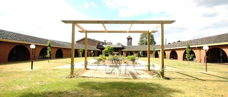 A courtyard with a pergola, a central table, and chairs. There are brick buildings with arched windows in the background.