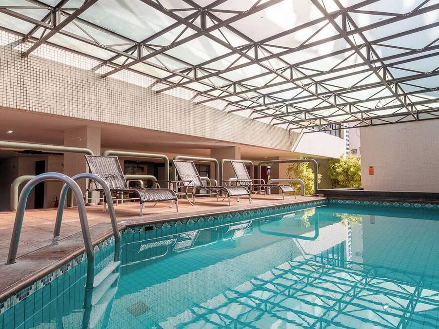 Piscina interna com teto de vidro, cadeiras de descanso e piso em azulejos.