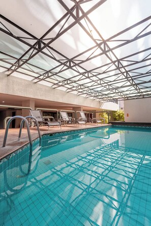 An indoor swimming pool with a glass ceiling, lounge chairs, and a tiled floor.