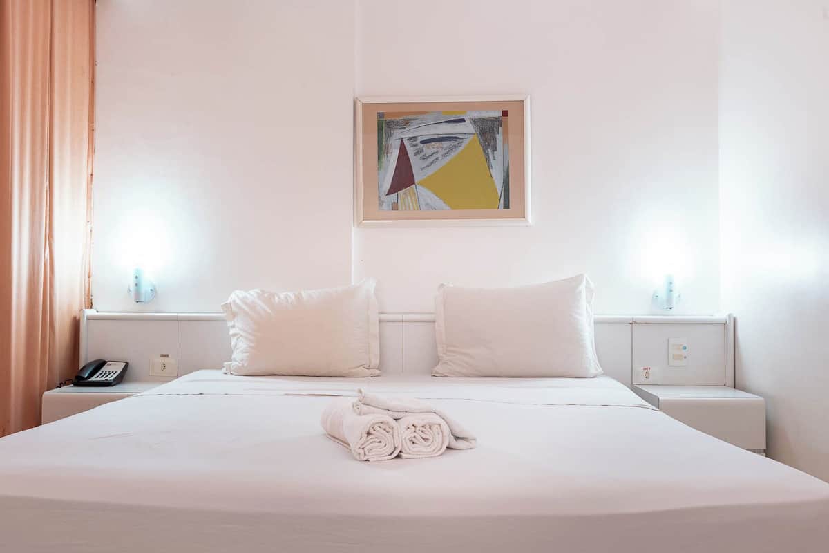 A hotel room with a large bed, two white pillows, a rolled towel on the bed, a painting on the wall, and bedside lamps.
