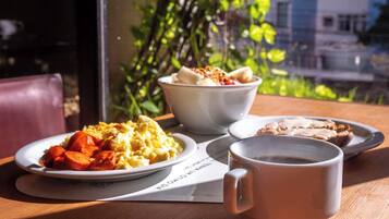 Una mesa con alimentos para el desayuno, incluyendo huevos revueltos, un tazón de cereal, tostadas y una taza de café, frente a una ventana con vistas a edificios y árboles.