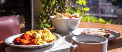 Una mesa con alimentos para el desayuno, incluyendo huevos revueltos, un tazón de cereal, tostadas y una taza de café, frente a una ventana con vistas a edificios y árboles.