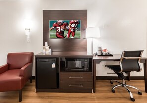 A hotel room with a desk, a red leather chair, a black office chair, a microwave, a mini-fridge, a lamp, and a sports-themed artwork on the wall.