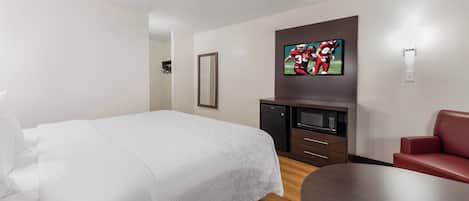A hotel room with a bed, a TV mounted on the wall, a small kitchenette, and a seating area with a table and chair.